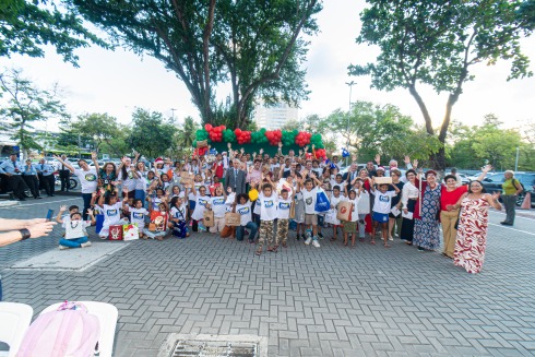 Grupo grande de crianças e adultos sorrindo ao ar livre, comemorando sob arco de balões.