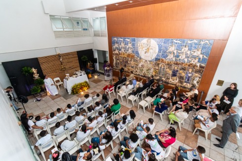 Crianças e adultos sentados em missa indoor com altar e grande painel artístico ao fundo.