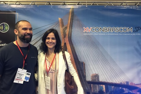 Fotografia de duas pessoas, na frente de um banner onde se lê &quot;Conbrascom&quot;