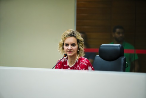 Foto mostra uma mulher adulta, loira, sentada e falando ao microfone