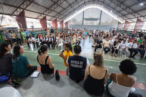 Na foto uma mulher fala para dezenas de pessoas sentadas numa quadra de esportes