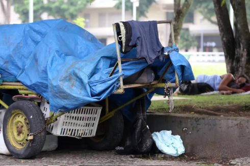 Foto mostra uma pessoa em situação de rua deitada no chão e uma carroça junto dela