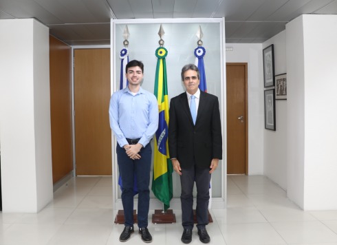 Duas pessoas posam em frente às bandeiras do Brasil e da Justiça do Trabalho em ambiente interno.
