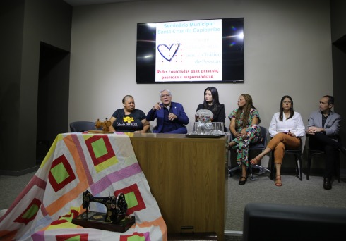 fotografia do desembargador Paulo Alcantara falando ao microfone. Ele se encontra no palco de um auditório e, junto com ele, há outras cinco autoridades