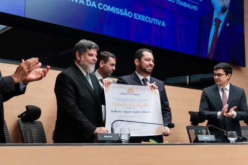 Homens de terno exibem diploma em cerimônia oficial, enquanto público aplaude.