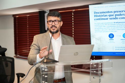 foto homem paletó falando ao púlpito; ao fundo, monitor de tv