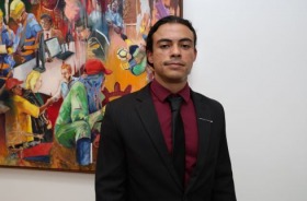 foto homem em pé vestindo terno preto; ao fundo quadro colorido