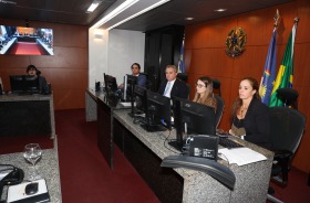 Foto mostra dois homens e duas mulheres sentados atrás de uma mesa de granito preto. Atrás deles, está o brasão da república