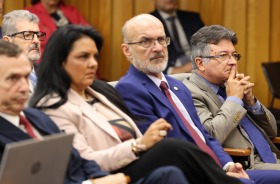 Foto mostra cinco pessoas sentadas em cadeiras de um auditório