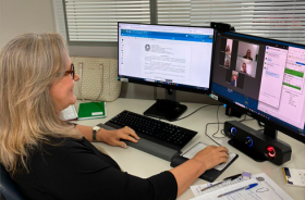 Foto onde aparece uma mulher diante de duas telas de computador, participando de uma tele audiência