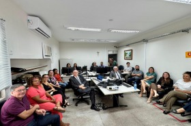 Grupo formado por 18 adultos sentados, reunidos numa sala de audiência