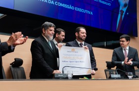 Homens de terno exibem diploma em cerimônia oficial, enquanto público aplaude
