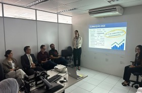 Na foto, uma mulher em pé, fazendo uma apresentação, e 5 adultos sentados