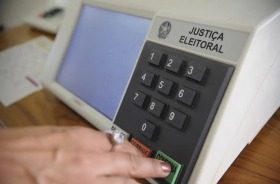 Fotografia da urna eletrônica brasileira e uma mão apertando a tecla 'confirma'