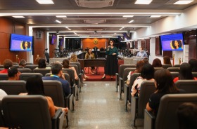 Foto mostra o auditório com pessoas sentadas assistindo à palestra de uma mulher