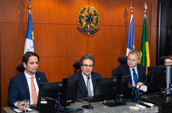 Foto mostra três homens adultos sentados em uma mesa, lado a lado, enquanto um fala ao microfone