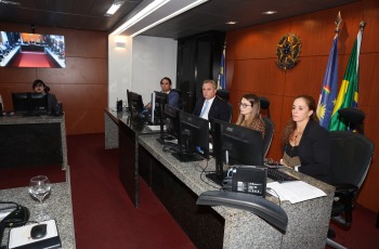 Foto mostra dois homens e duas mulheres sentados atrás de uma mesa de granito preto. Atrás deles, está o brasão da república