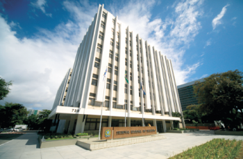 Foto mostra a fachada do TRT 6, com grama no canto inferior direito e um ceu azul ao fundo