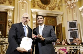 foto dois homens em pé segurando certificado e medalha em salão nobre